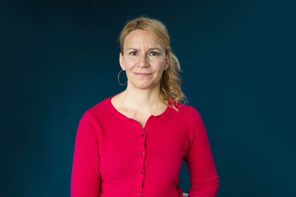 Punaiseen puseroon ja rengaskorviksiin pukeutunut Kaari Mattila seisoo vihreäsävyisen seinän edessä ja katsoo kameraan.