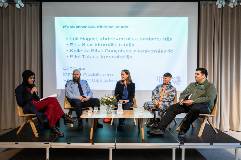 Lavalla istuu kaaressa viisi keskustelijaa. Keskellä on pöytä ja taustalla valkokangas.