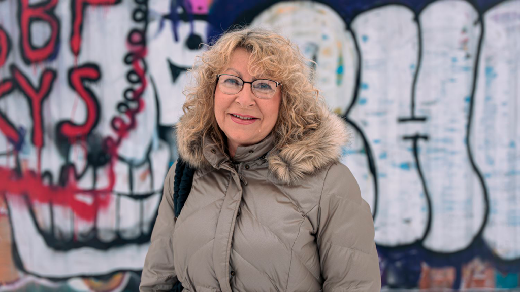 Virpi katsoo suoraan kameraan varovasti hymyillen. Taustalla näkyy graffitiseinä.