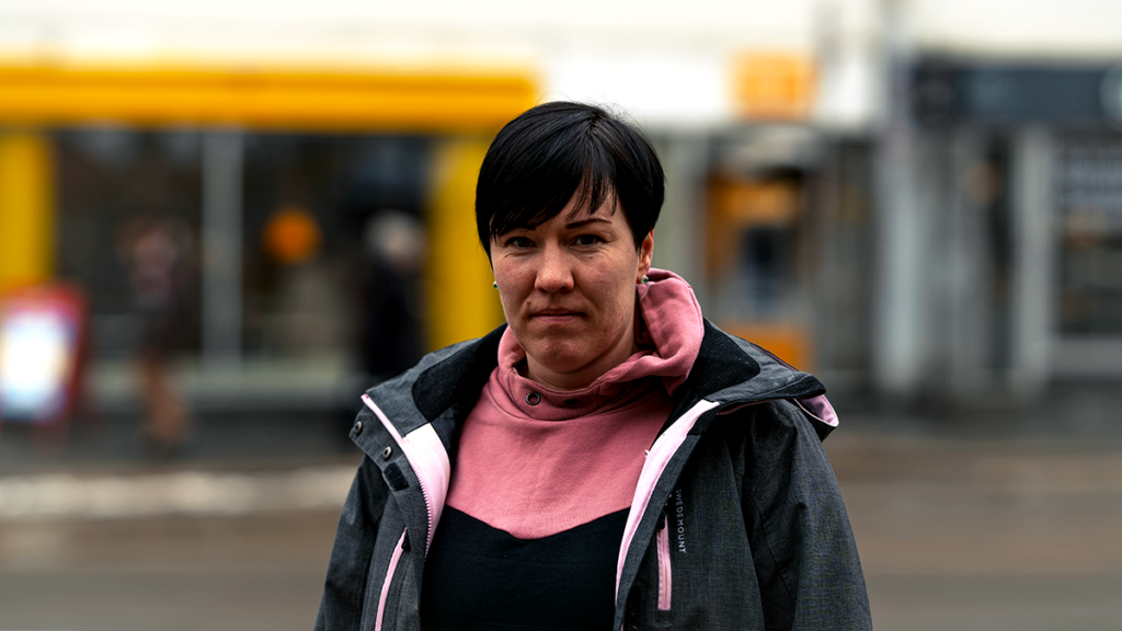 Maria katsoo vakavana kameraan. Taustalla bussi ja kaupunkinäkymää.