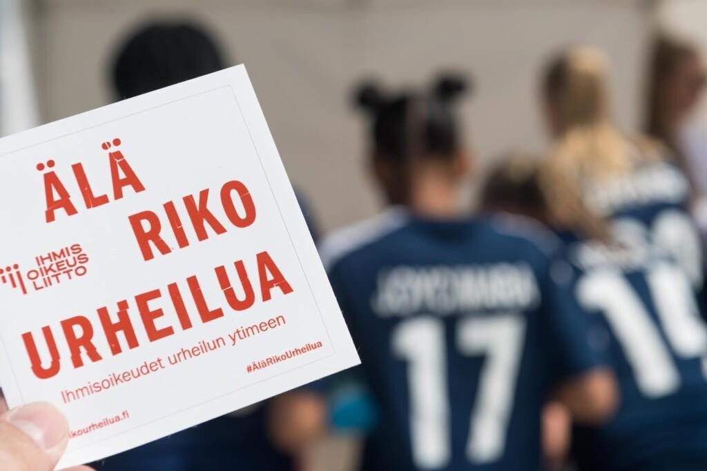 Etualalla tarra, jossa lukee "Älä riko urheilua. Ihmisoikeudet urheilun ytimeen". Taustalla sumeana peliasuihin pukeutuneita urheilijoita.